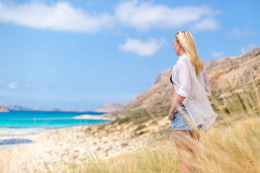 Relaxed woman in white shirt looking at distance, enjoying beautiful nature, freedom and life at serene landscape at Balos beach, Greece. Concept of vacations, freedom, happiness, joy and well being.