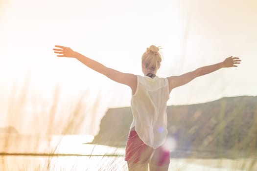 Relaxed woman in white shirt, arms rised, enjoying sun, freedom and life an beautiful beach. Young lady feeling free, relaxed and happy. Concept of vacations, freedom, happiness, joy and well being.