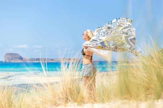 Relaxed woman, arms rised, holding colorful scarf, enjoying sun, freedom and life at beautiful beach. Young lady feeling free, relaxed and happy. Concept of vacations, freedom, joy and well being.