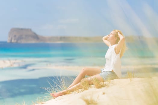 Relaxed woman enjoying sun, freedom and life an a beautiful sandy beach of Balos in Greece. Young lady feeling free, relaxed and happy. Vacations, freedom, happiness, enjoyment and well being.