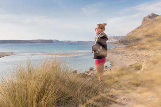 Relaxed woman in dark hoodie and colorful scarf looking at distance, enjoying beautiful nature, freedom and life at serene landscape at Balos beach, Greece. Concept of well being.