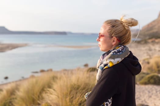 Relaxed woman in dark hoodie and colorful scarf looking at distance, enjoying beautiful nature, freedom and life at serene landscape at Balos beach, Greece. Concept of well being.