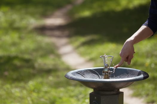 Horizontal close up of a hand pushing a metal water dispenser in a green park