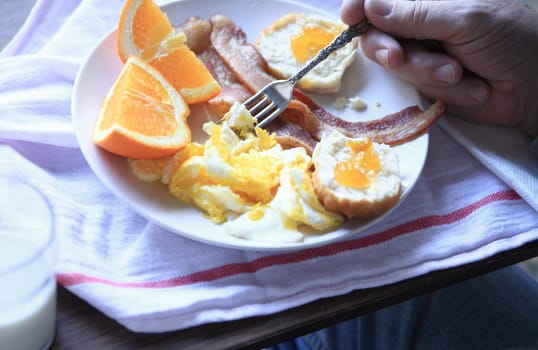 A man eats a hearty breakfast of bacon, scrambled eggs, biscuit with jam and orange sections.