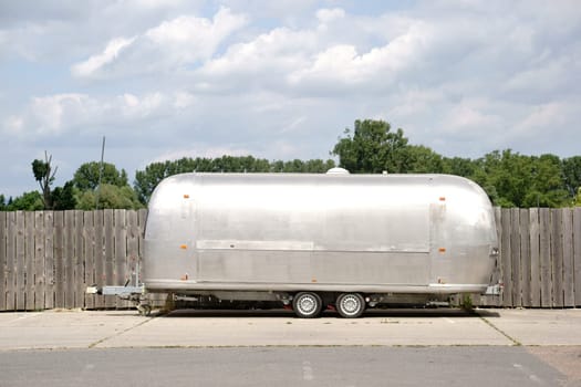A round silver-colored trailer made of sheet metal stands in a parking lot with a board fence.