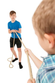 Two brothers pull each at the ends of a rope isolated on white. Younger brother (9 years) blurred in the foreground with his back to the camera. Older brother (13 years) sharply depicted in the background as a full-body view facing the camera.