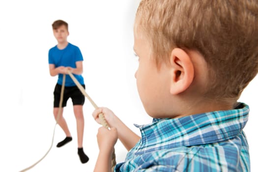 Two brothers pull each at the ends of a rope isolated on white. Younger brother (9 years) sharp in the foreground with his back to the camera. Older brother (13 years) blurred in the background as a full-body view facing the camera.