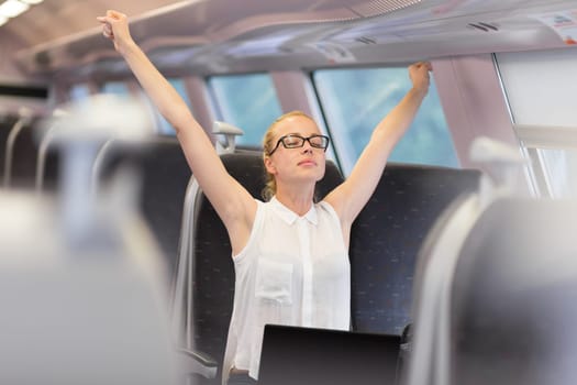 Businesswoman traveling by train, streching her upper body arms reised taking a break from laptop work. Business travel concept.