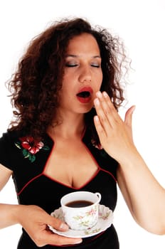 A really tired woman in a black dress holding the hand over her mouth,.yawning, with a cup of coffee, isolated for white background..