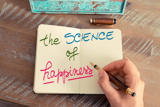 Retro effect and toned image of a woman hand writing a note with a fountain pen on a notebook. Handwritten text The Science of Happiness as success and evolution concept image