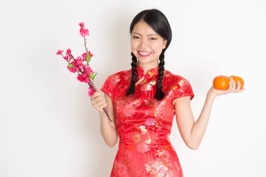 Portrait of Asian Chinese girl hands holding tangerine orange and plum blossom, in traditional red cheongsam standing on plain background.