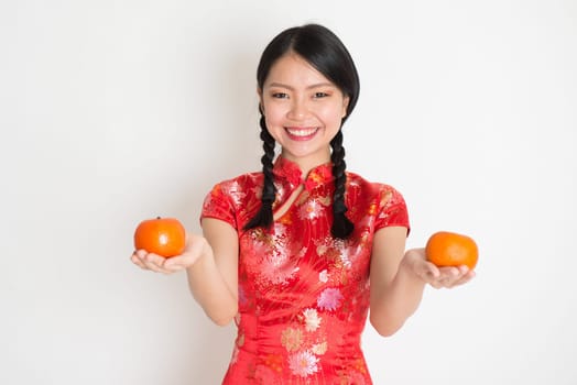 Portrait of Asian Chinese girl hands holding tangerine fruit, in traditional red qipao standing on plain background.