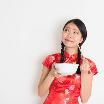 Asian Chinese female eating using chopsticks holding rice bowl, in traditional cheongsam on plain background, thinking and looking up at copy space.