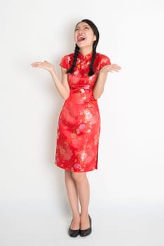 Portrait of full body Asian Chinese girl with excited facial expression, open palms and looking up, in traditional red qipao standing on plain background.