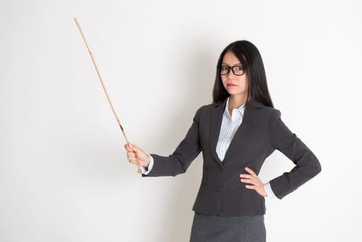 Asian female teacher angry and holding a stick, standing on plain background.