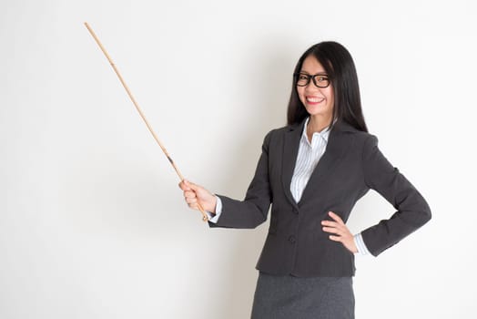 Asian female teacher smiling and holding a stick, standing on plain background.