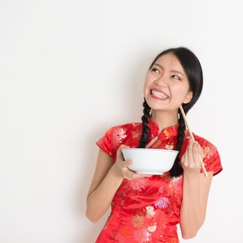 Asian Chinese female eating using chopsticks holding rice bowl, in traditional qipao on plain background, thinking and looking up at copy space.