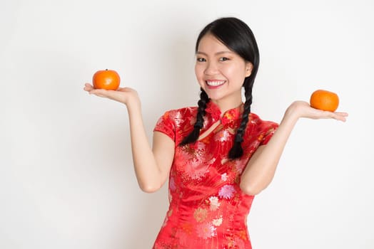 Portrait of Asian Chinese girl hands holding tangerine orange, in traditional red qipao standing on plain background.