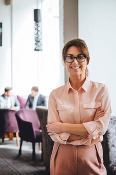 Business woman standing with her arms folded. She is smiling at the camera. Two men in suits can be seen talking in the background.