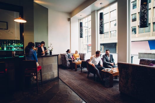 Full room shot of an active hotel bar and cafe. There are people sitting and drinking. Two business men are discussing work.