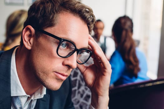Close up of a businessman looking stressed. He has his hand on his head and there are other people in the background.