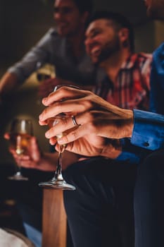 Close up of a mans hands holding a glass of wine in a bar. There are two men laughing in the background.