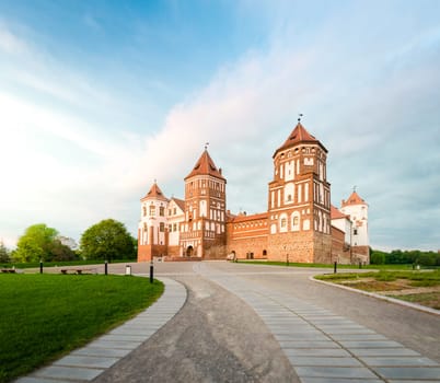Green trees and gras and blue sky at sunny day. Stone road to beautiful Mir castle in Belarus. Medieval fort became sight for tourists.