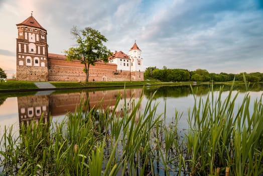 Green trees and gras, blue sky and lake at sunny day. Beautiful reflection of Mir castle on water. Medieval fort became sight for tourists in Belarus.