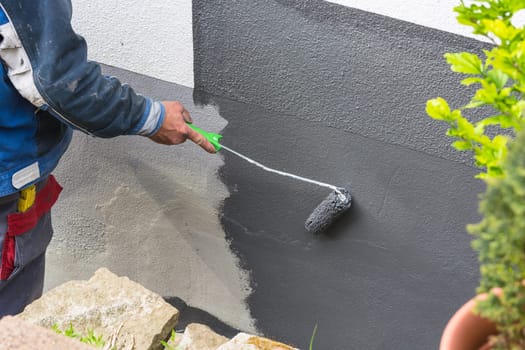 Close-up arm of a painter with paint roll in his hand paints a house wall with gray paint.
