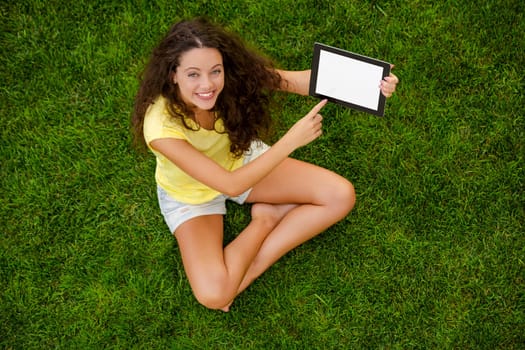Beautiful young woman sitting on the grass and holding a tablet