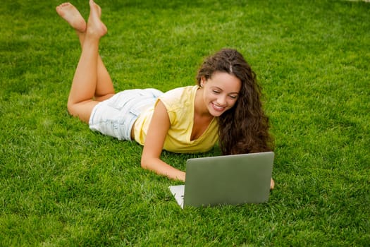 Beautiful young woman lying on the grass and working with a laptop