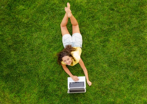 Beautiful young woman lying on the grass and working with a laptop