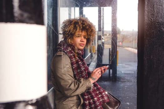 Woman using her smart phone as she waits for her train at the platform. She is sitting in the shelter, looking out for the train.