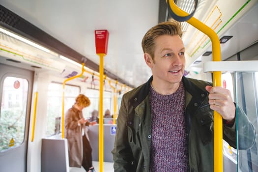 Man standing on the train holding on to the hand rail. He is looking out the window with a cheerful expression.