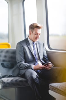 Formal business man sitting on the train with his bag. He has a smart phone in his hands, which he is looking down at.