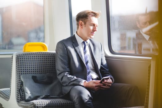 Formal business man sitting on the train with his bag. He is looking out the window and has a smart phone in his hands.