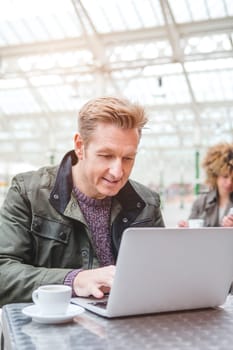 Mature man sitting at a table outside of a cafe in a train station. He is enjoying a cup of tea while he uses his laptop.