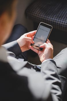 Over the shoulder view of a formal businessman sitting on a train. He has a smart phone in his hands, which has information about a meeting on the screen.