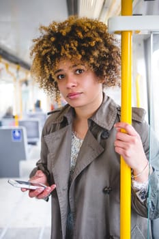Woman commuting on the train. She is holding on to the handrail and has her smart phone in the other hand, while looking at the camera.