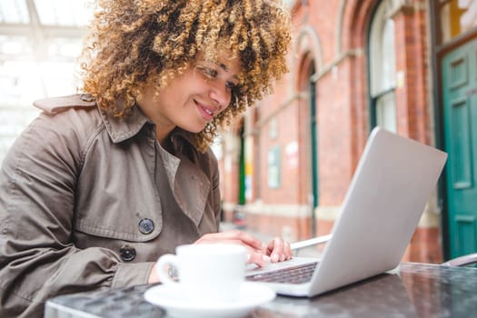Woman using a laptop in a cafe. She is sat outside at a table with a coffee.