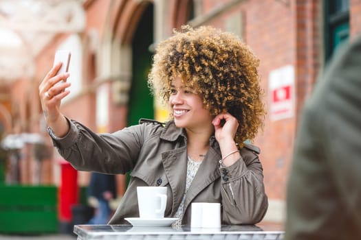 Woman taking a selfie on a smartphone. She is sat outside a cafe and is using a smart phone to take a photo.
