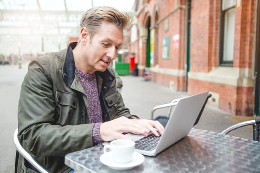 Man using a laptop at a cafe. He is sat at a table outside with a coffee.