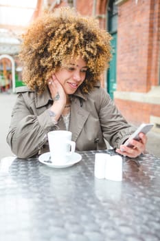 Woman sitting outdoors at a coffee shop with a drink. She is using a smart phone.