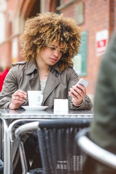 Woman sitting outdoors at a coffee shop with a drink. She is using a smart phone.