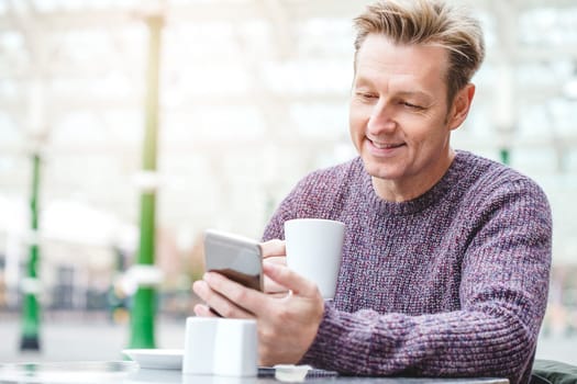 Man enjoying a hot drink at a cafe in a train station. He is sitting outside, looking down at his smart phone which is in his hand.
