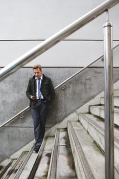 Businessman stood against the wall on a staircase. He is using a smart phone.