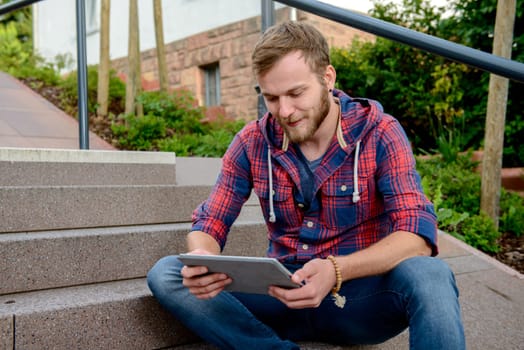 young man in checkered shirt sitting on stairs and using tablet PC