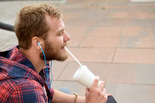 young bearded man in checkered shirt sitting on stairs listening music and drinking soft drink