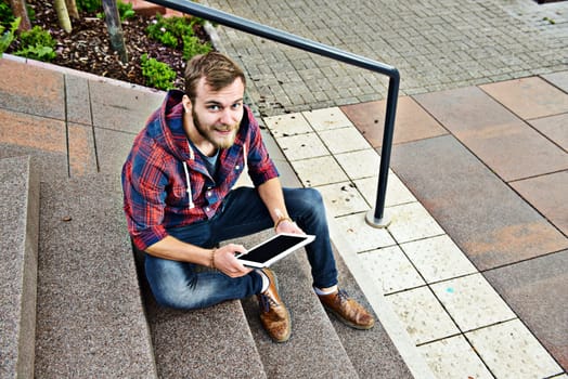 young man in checkered shirt sitting on stairs and using tablet PC