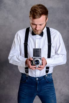 young bearded man in white shirt and bow tie with old camera standing in front of gray background